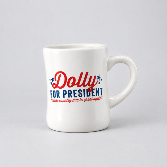 A white ceramic mug with the text 'Dolly FOR PRESIDENT' in red and blue, subheading: make country music great again