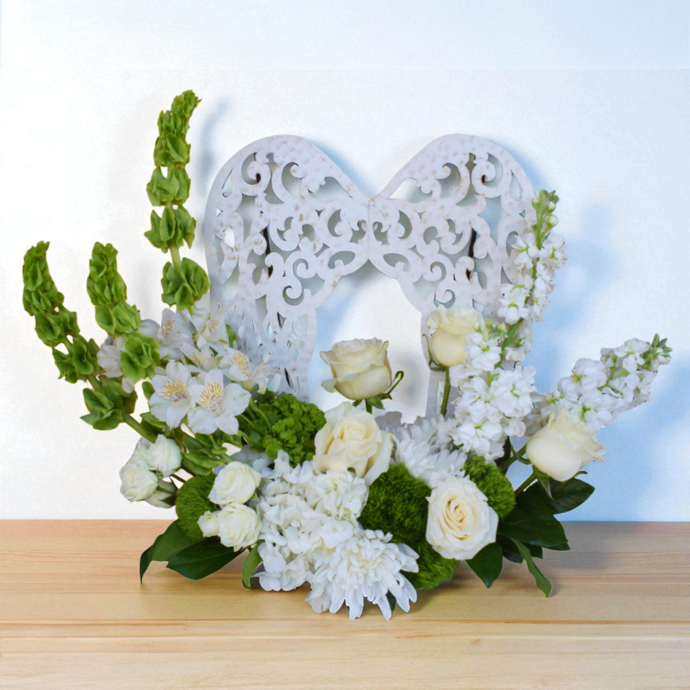 Angel’s Embrace is a large sympathy arrangement with white and green blooms framing a filigree metal angel wing keepsake, symbolizing protection and everlasting love.
